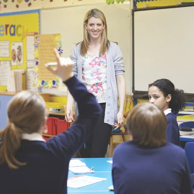 Teacher standing at front of class, looking at student in foreground with raised hand